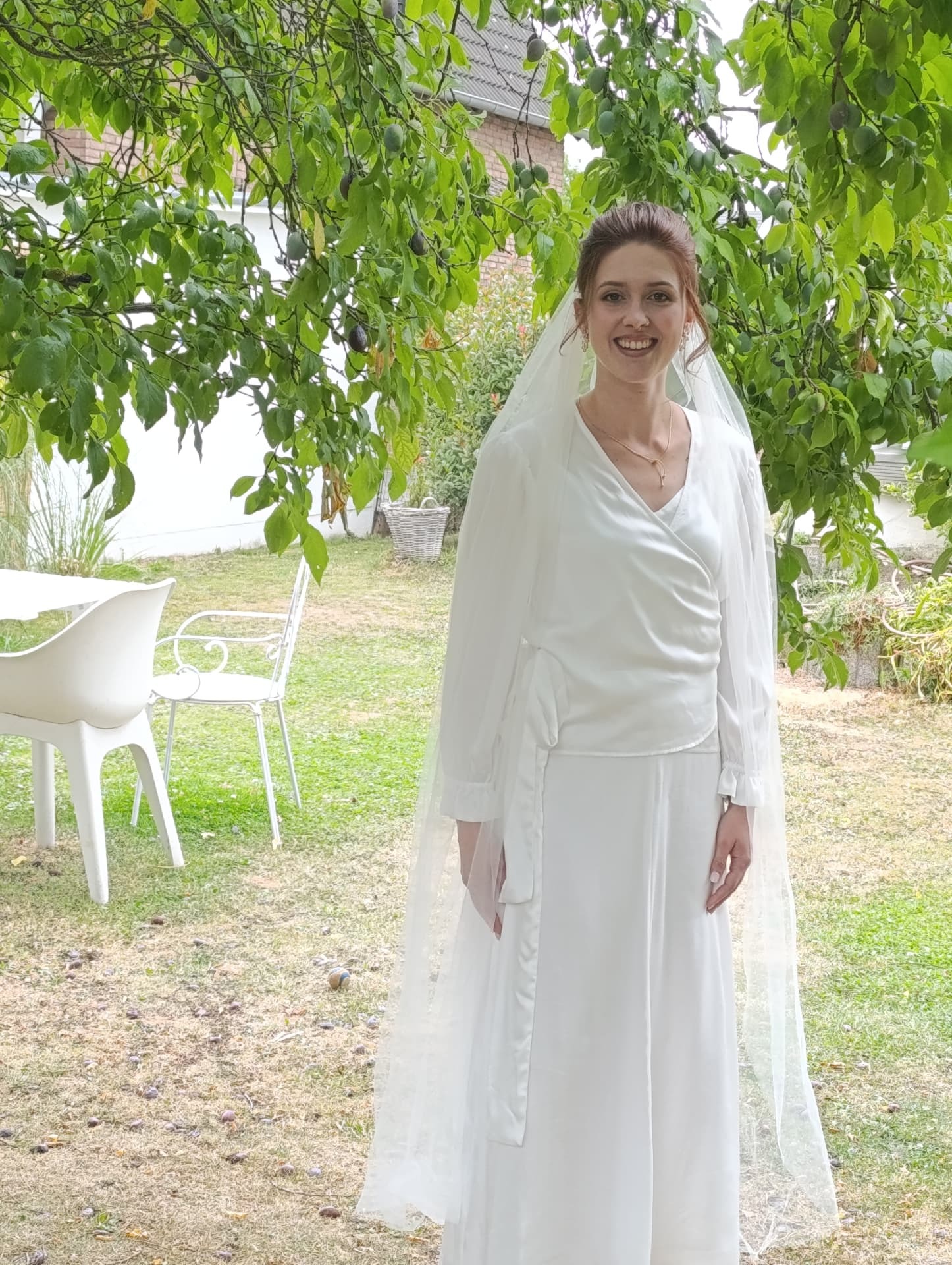 Braut im bodenlangen Brautensemble mit langem Tüllschleier, die in einem Garten lächelnd in die Kamera blickt