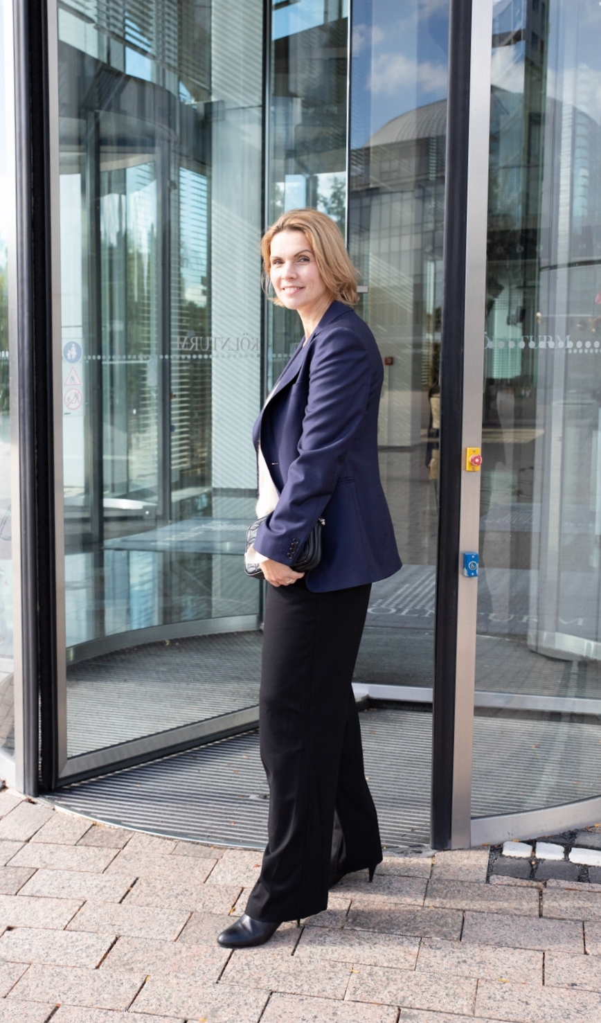 Frau in dunkelblauem Blazer und schwarzer Hose vor einer modernen Glasfassade
